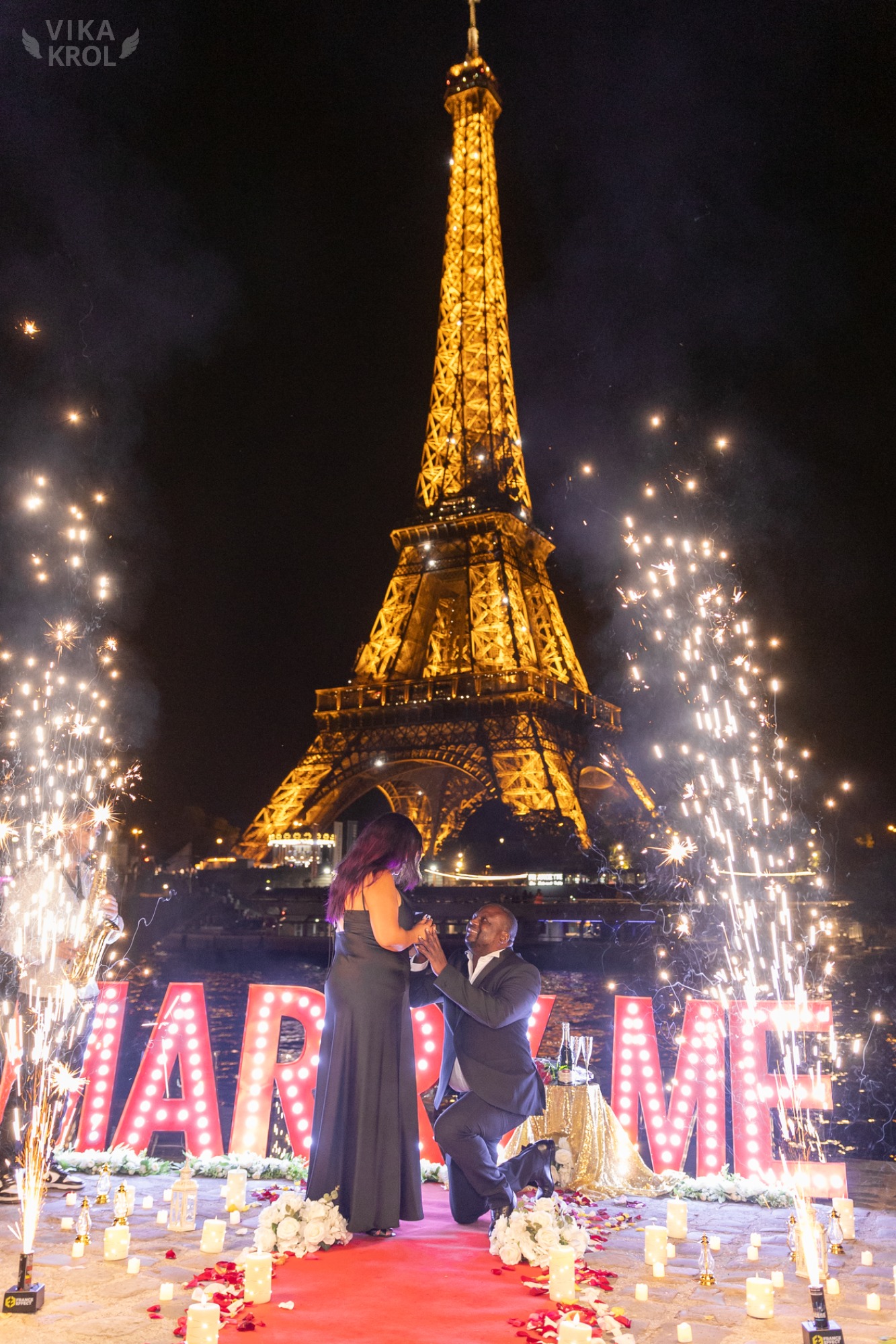 A couple stepping onto a Parisian rooftop. He leads his partner, who is blindfolded, toward a path lit by candles, with the Eiffel Tower glowing softly in the background.