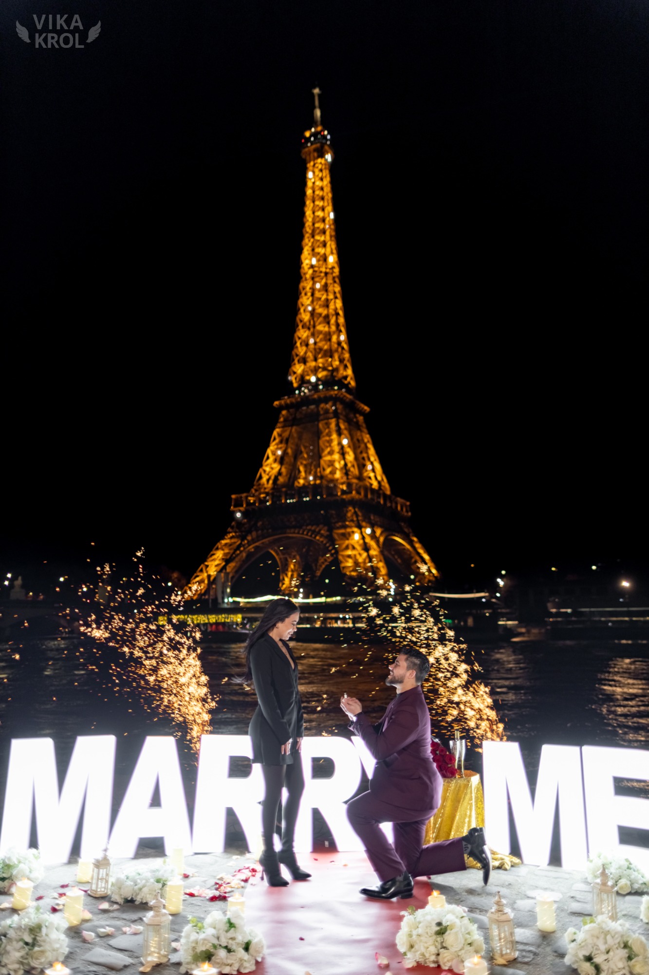 Una splendida, ampia inquadratura da un tetto privato parigino di notte. Una coppia è in un dolce abbraccio, con bicchieri di champagne in mano. La Torre Eiffel brilla intensamente sullo sfondo, apparendo perfettamente inquadrata e incredibilmente vicina. La scena è illuminata da decine di candele calde e lanterne a terra, con petali di rosa sparsi su un tappeto rosso scuro. Non c'è nessun altro in vista.